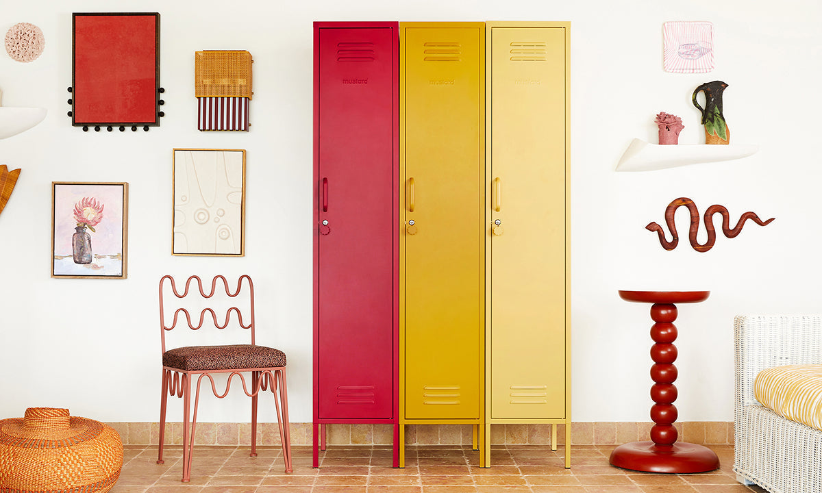 Les casiers « Skinny » aux couleurs Poppy, « Mustard » et Butter sont alignés contre un mur de galerie, accompagnés d’une chaise ondulée et d’une table d’appoint.