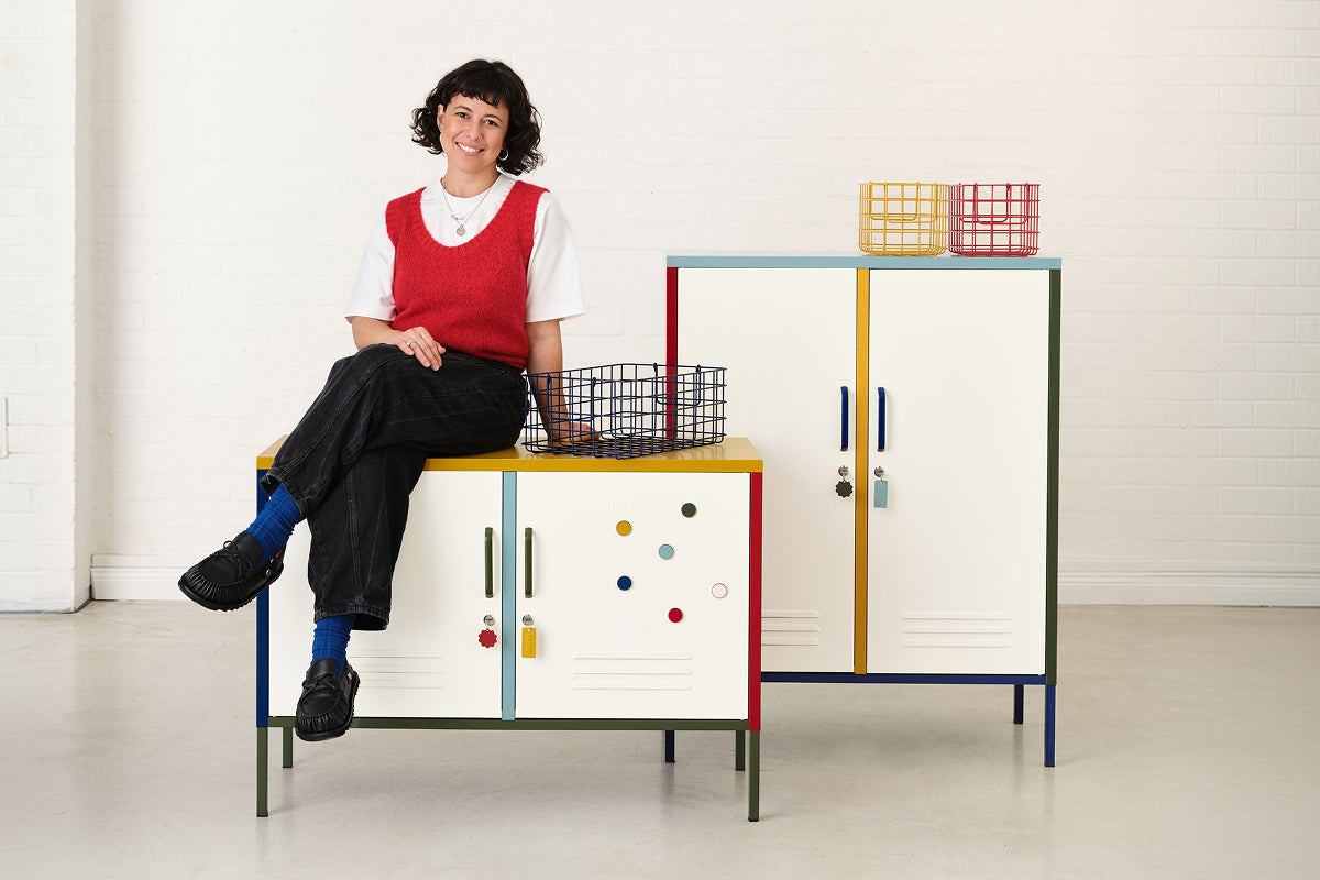 Becca sitzt auf dem „ Lowdown “-Spind neben dem „ Midi “, dekoriert mit Drahtkörben und „ Buttons “ in der Farbpalette „ Mustard x MoMA“