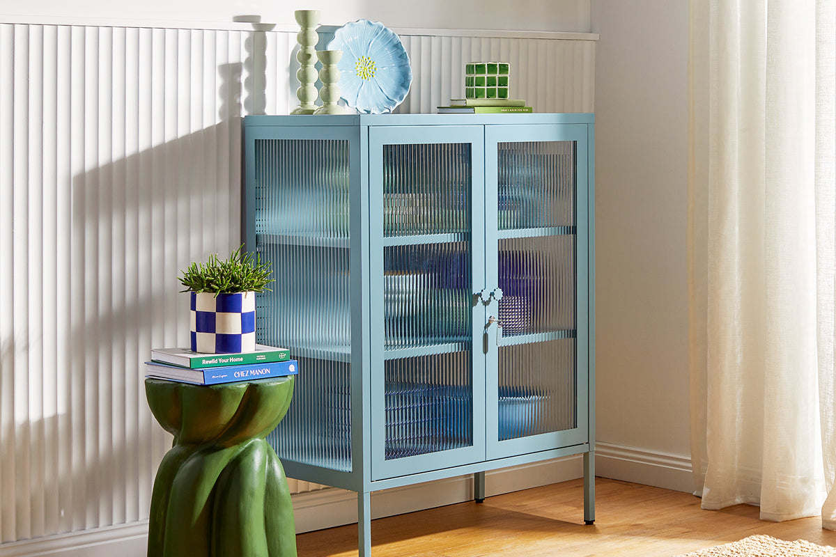 Mustard La vitrine « Mixer » de Made, dans la teinte « Ocean » (un bleu pâle et poussiéreux), mise en valeur par des objets en céramique posés devant des lambris cannelés.