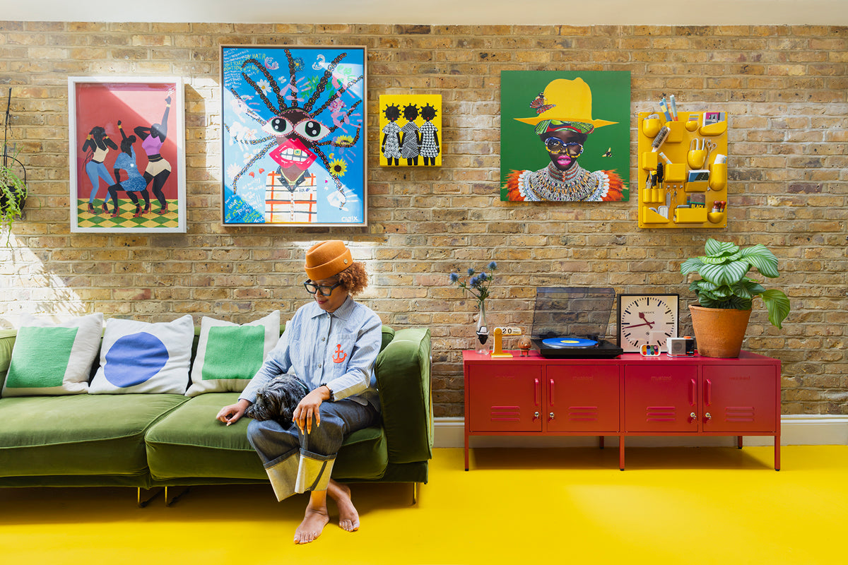 Natasha Landers, sentada en un sofá de terciopelo verde bajo una galería de obras de arte de colores colgadas en una pared de ladrillo, junto a unas taquillas rojas Poppy sobre un suelo amarillo