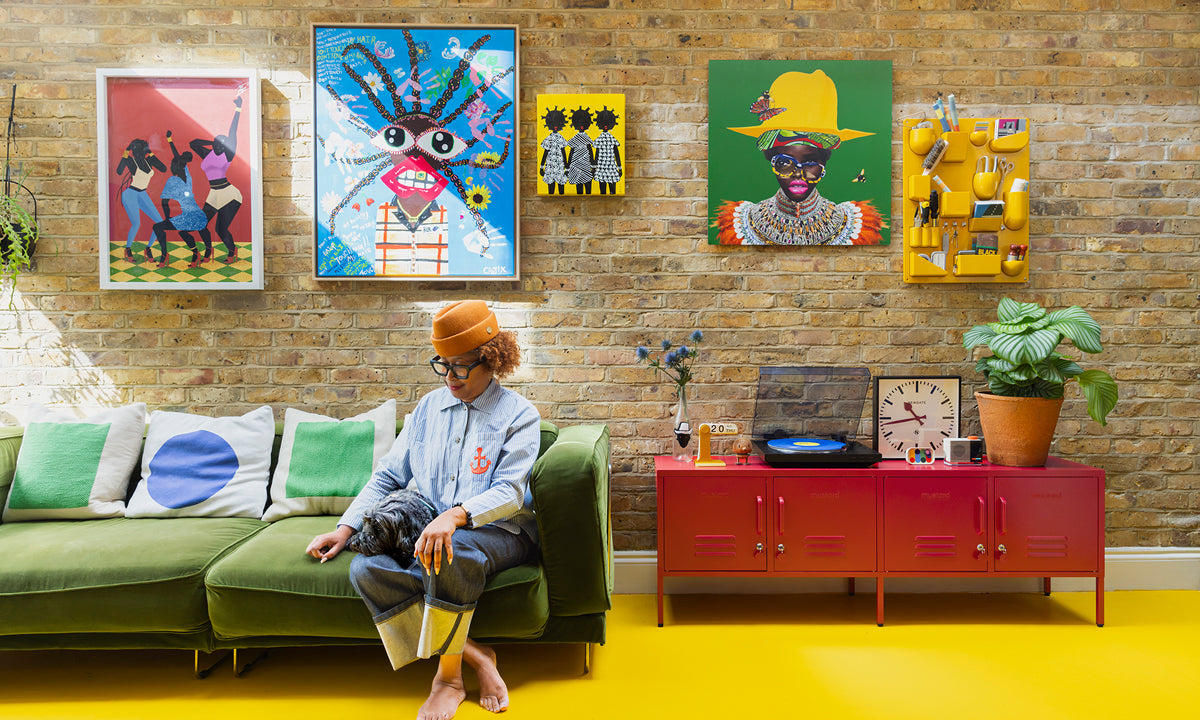 Natasha Landers on a green velvet sofa under a brick wall gallery of colourful art, beside Poppy red lockers on a yellow floor