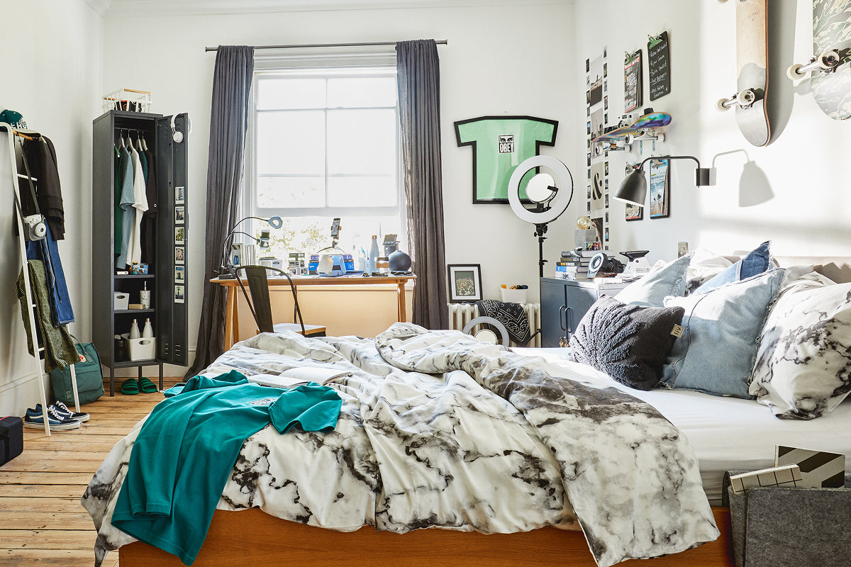 Der Skinny in der Farbe „Slate“, der als Kleiderschrank in einem Schlafzimmer einer Wohngemeinschaft dient, mit einem Schreibtisch am Fenster und Skateboards an der Wand