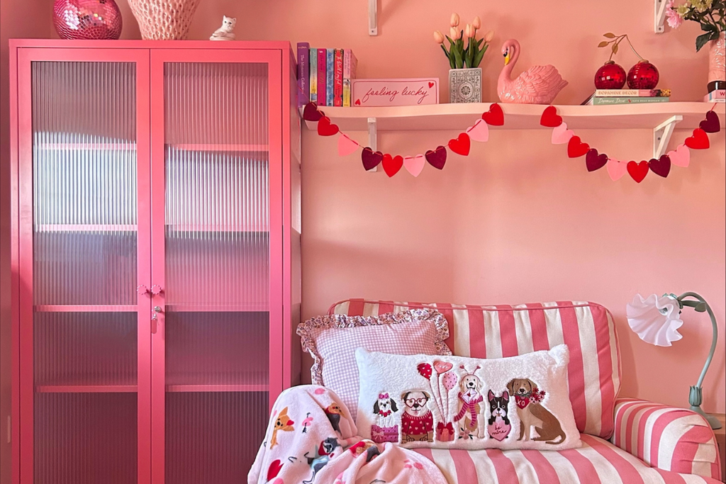 A pink-themed living room featuring a pink Mustard Made locker with fluted glass doors, a striped pink and white arm chair and two floating shelves displaying knick-knacks.