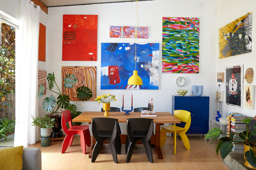 A colourful dining room featuring white brick walls, large abstract paintings and quirky chairs in red, black and yellow.