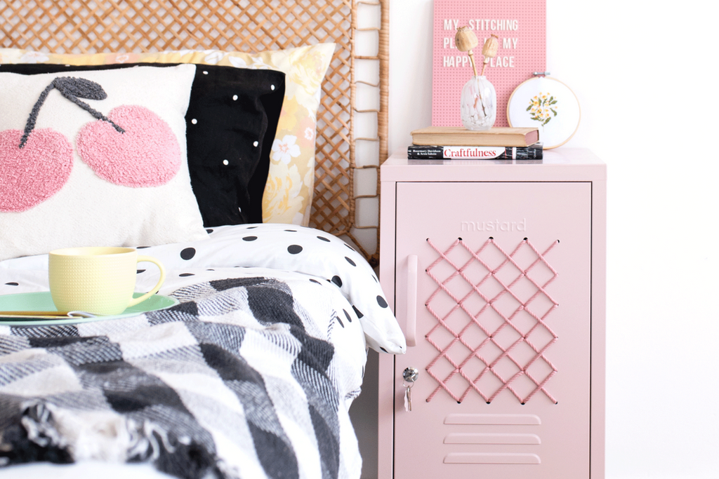 A short blush locker featuring a cross-stitch patterned door is styled next to a rattan bed with a mix of patterned linens and cushions.