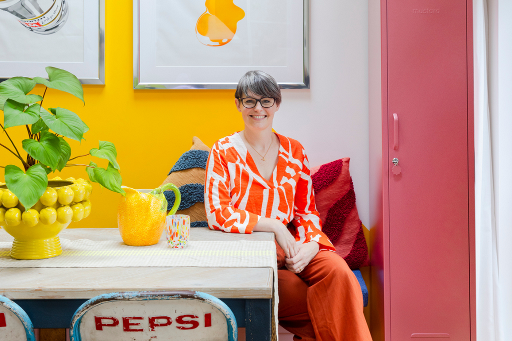 Genie is wearing a bright orange outfit and smiling as she sits at her dining table. There is a Berry Skinny locker next to her and a vibrant yellow wall behind.