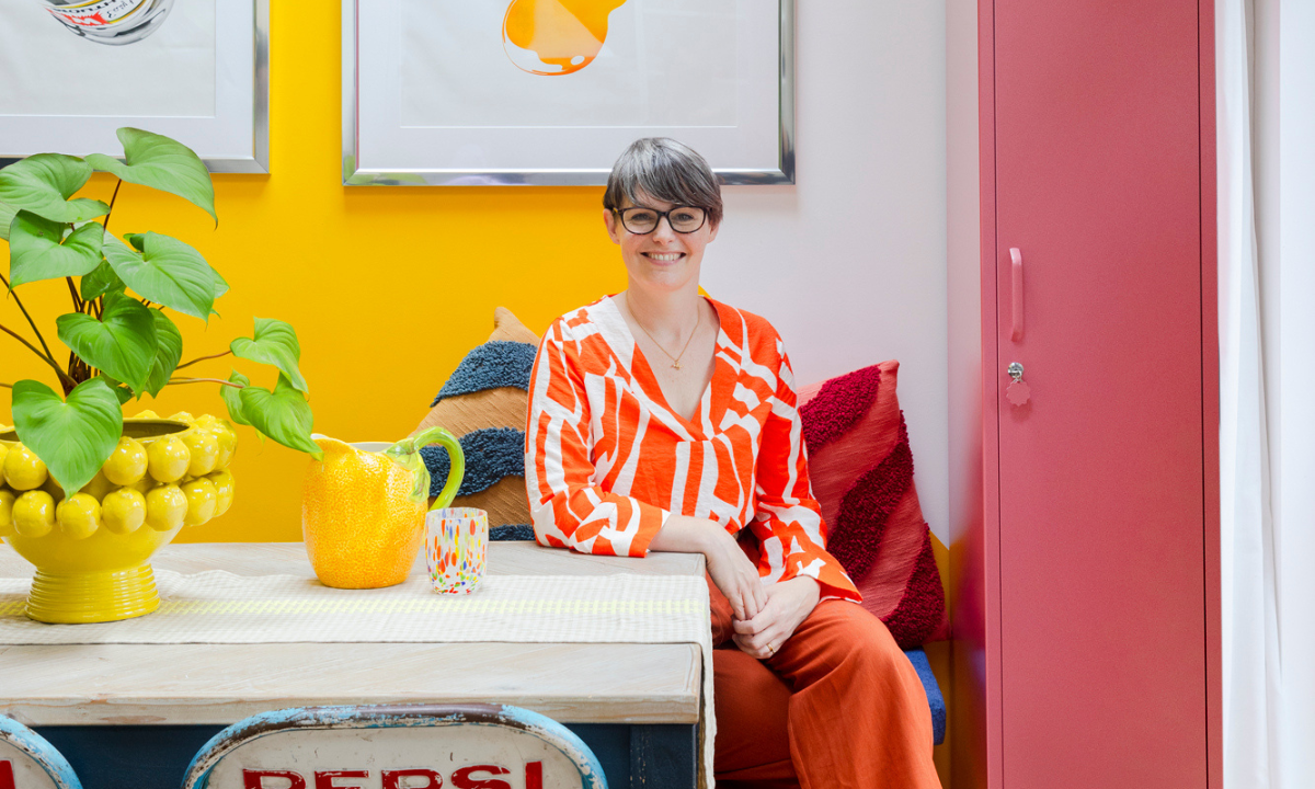 Genie is wearing a bright orange outfit and smiling as she sits at her dining table. There is a Berry Skinny locker next to her and a vibrant yellow wall behind.