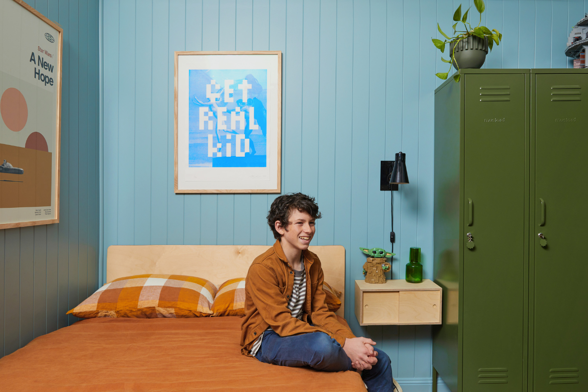 Dylan smiles as he sits on his bed. There is an Olive Twinny behind him and rust-coloured linens on the bed.