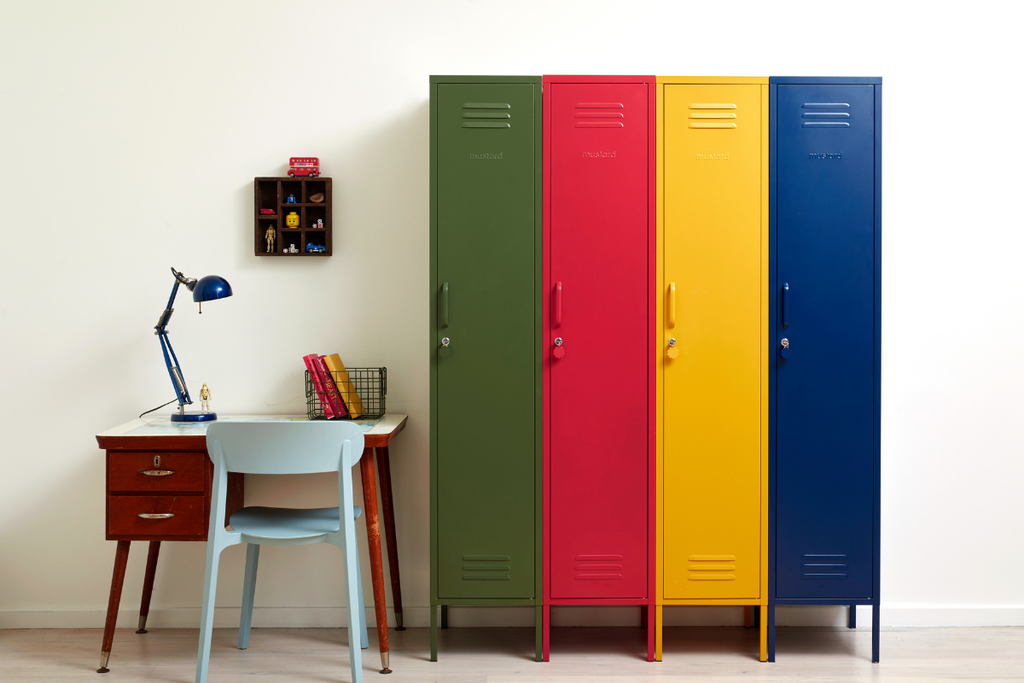 Four brightly coloured Skinny lockers sit side by side to create a rainbow of primary colours. Next to them is a small wooden desk with a pale blue chair.