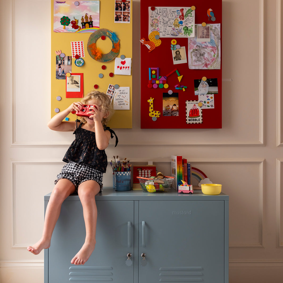 Child sitting on The Lowdown locker in Ocean blue below two Storyboard magnetic boards in Mustard yellow and Poppy red