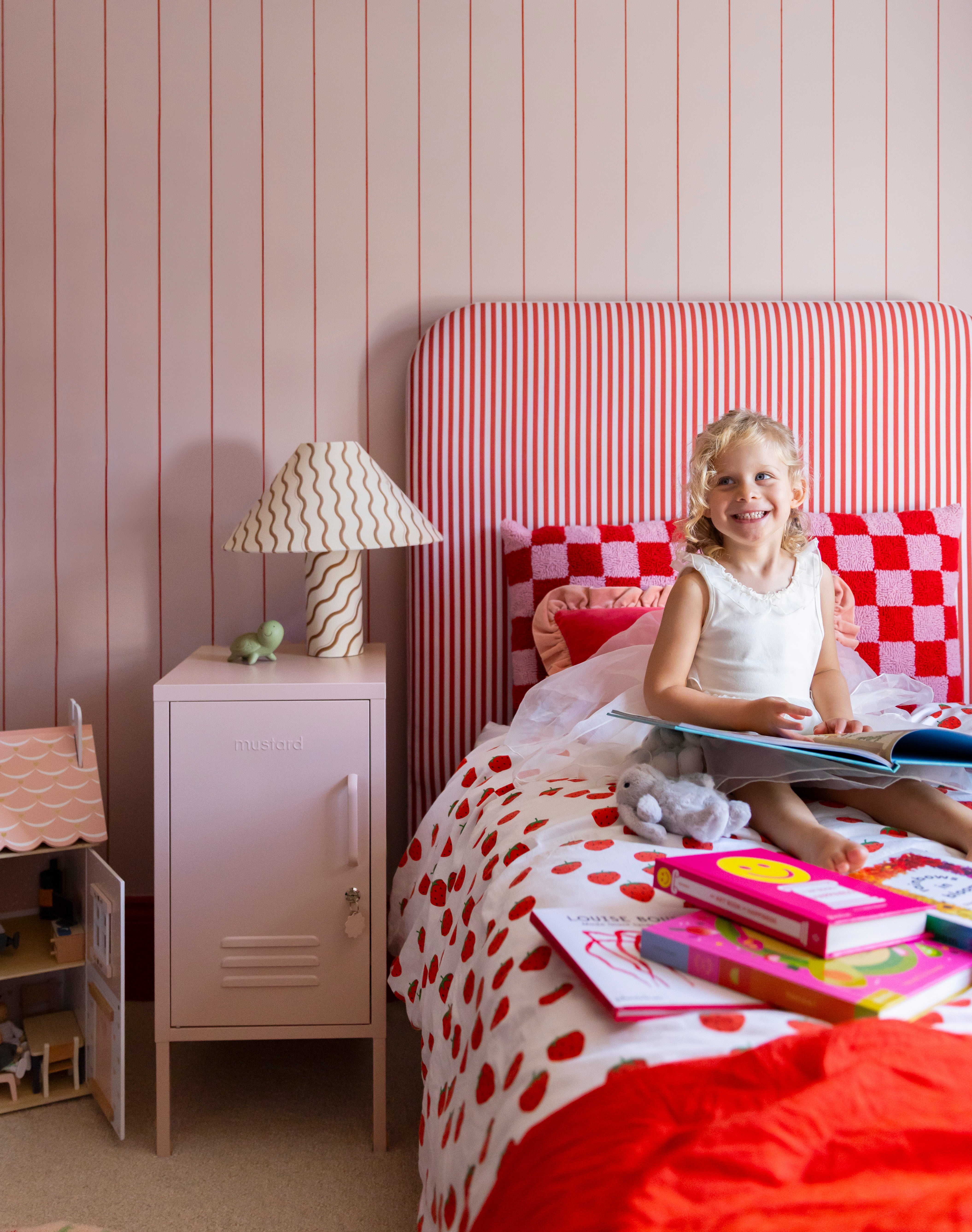 A young girl sits on a bed reading books in her pink and red styled bedroom, with a Blush pink locker as her bedside table.