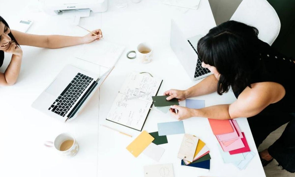 Becca beugt sich über einen weißen Tisch, um Jess Farbmuster zu zeigen