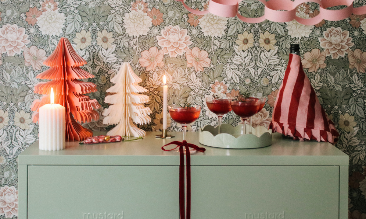 Die Oberseite eines Schranks von Sage Mustard Made ist mit rotem und rosa Weihnachtsschmuck und brennenden Kerzen dekoriert. Dahinter befindet sich eine rosa und grüne Blumentapete mit einer rosa Papierkette.