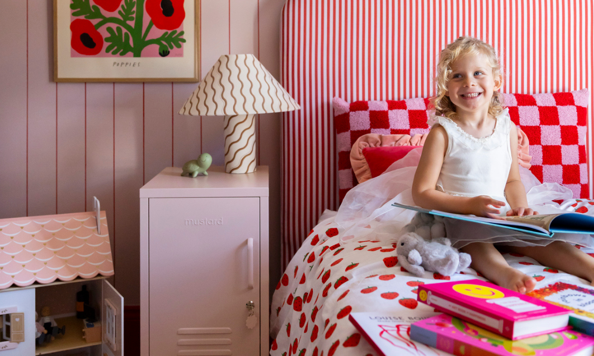 Una niña está sentada en una cama leyendo libros en su dormitorio de estilo rosa y rojo, con una taquilla rosa rubor como mesilla de noche.