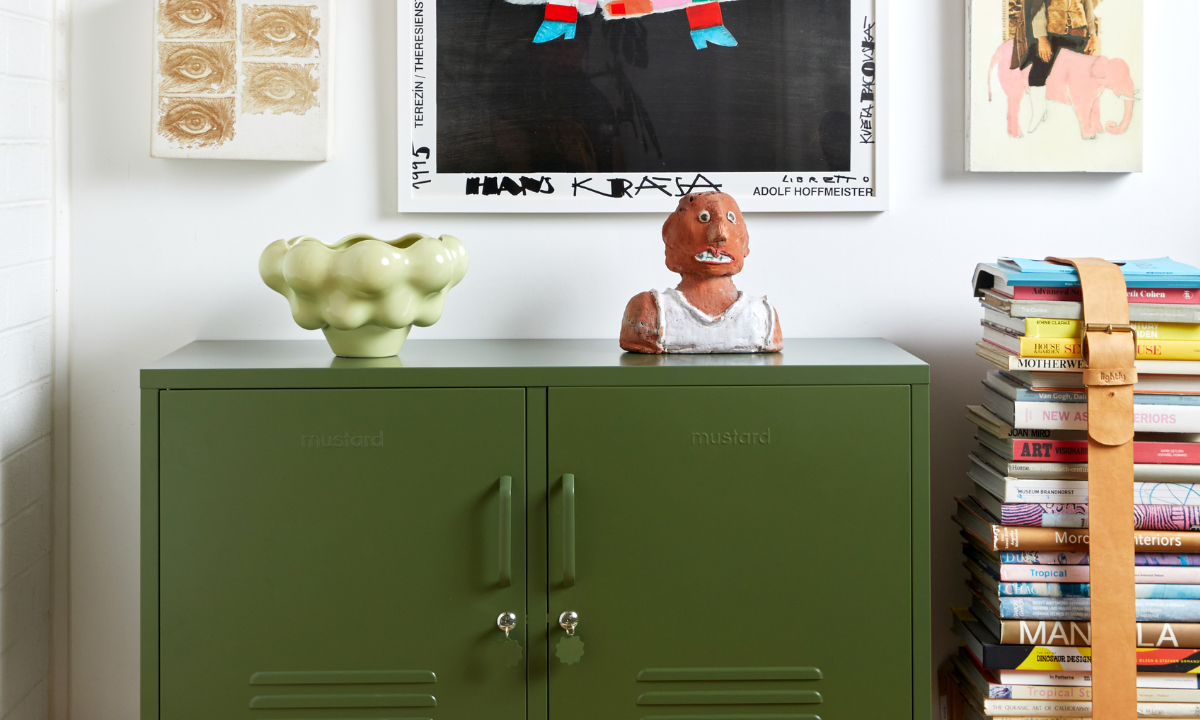 Un armario Olive Lowdown Mustard Made Locker junto a una pila de libros de sobremesa y una ecléctica gama de coloridas obras de arte.