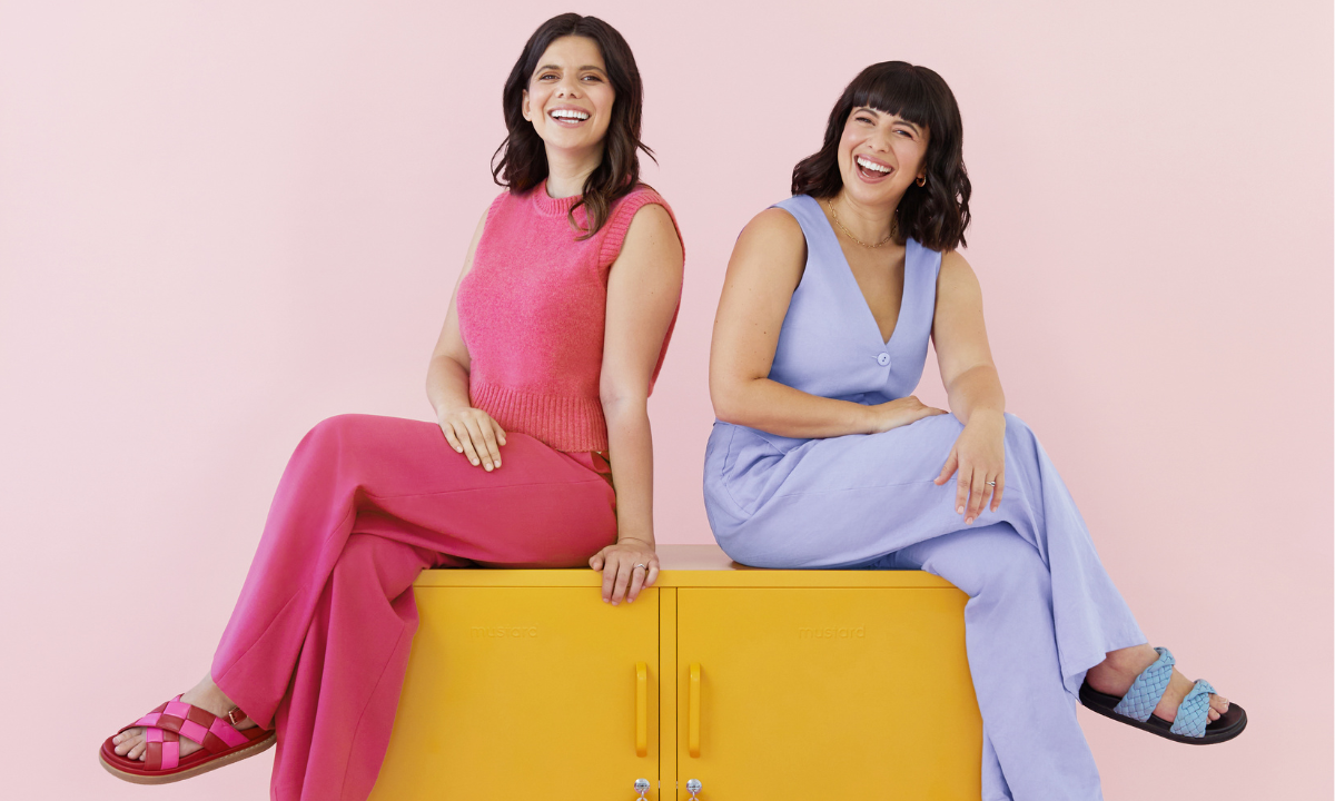 Becca y Jess se sientan en The Lowdown in Mustard. Jess lleva una camiseta de punto de color rosa baya y pantalones largos. Becca lleva un chaleco morado lila claro y pantalones largos. 