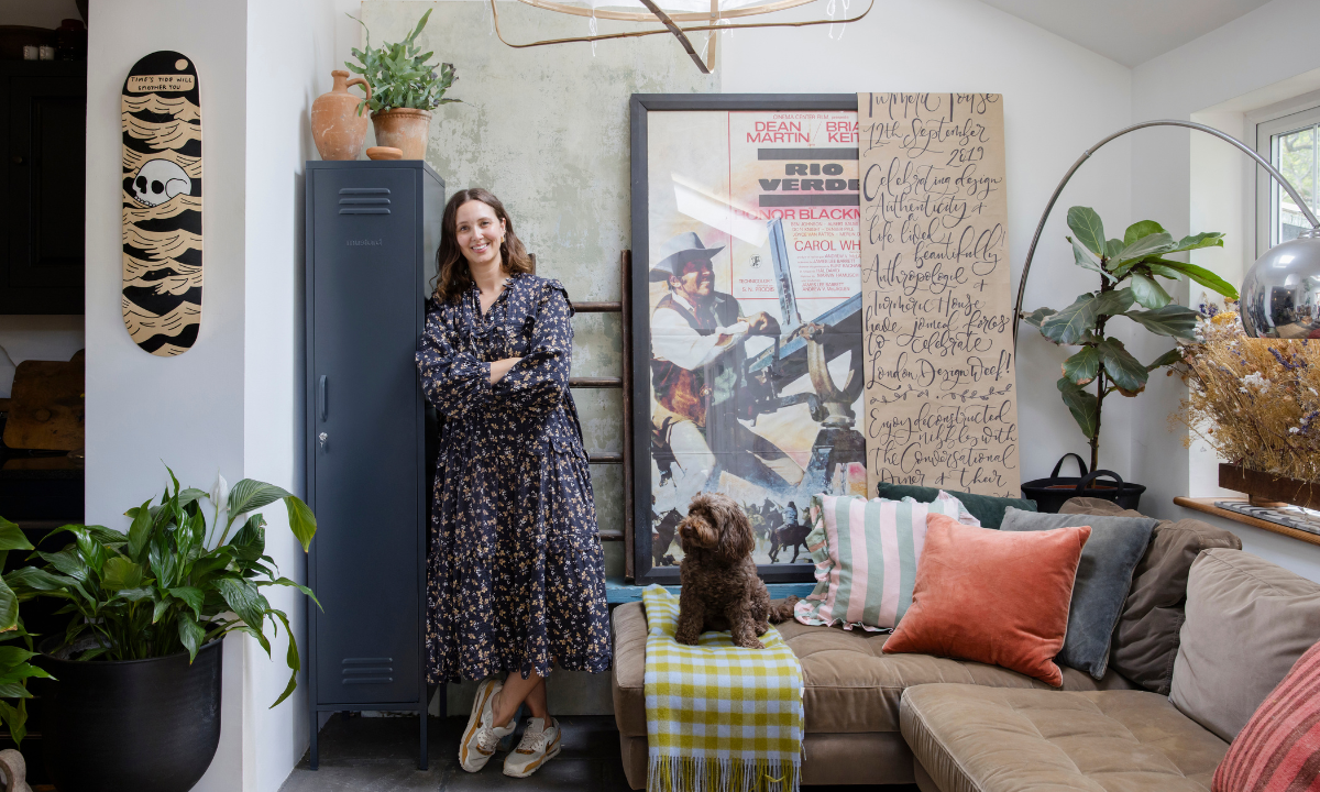 Clare Watson lleva un vaporoso vestido de flores y zapatillas deportivas mientras se apoya en una taquilla Skinny Mustard Made en el salón de su casa. Está rodeada de plantas, obras de arte, cojines y un perrito en el sofá.
