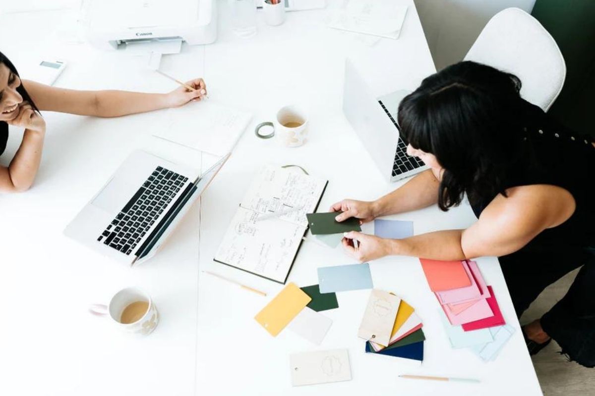 Becca se penche sur une table blanche pour montrer à Jess des échantillons de couleurs.