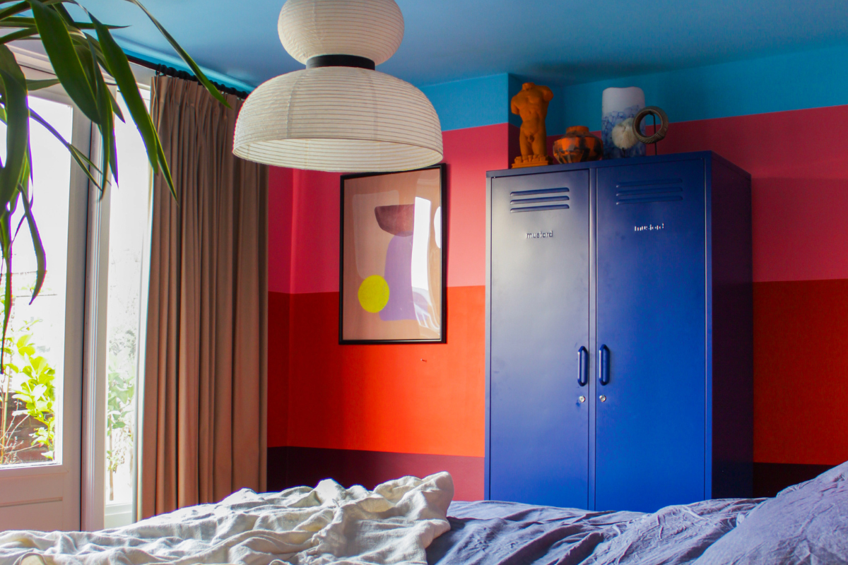 Una camera da letto è dipinta con spesse strisce orizzontali di prugna, rosso e rosa, con un soffitto azzurro. C'è un armadietto Navy Twinny accanto a una stampa astratta.